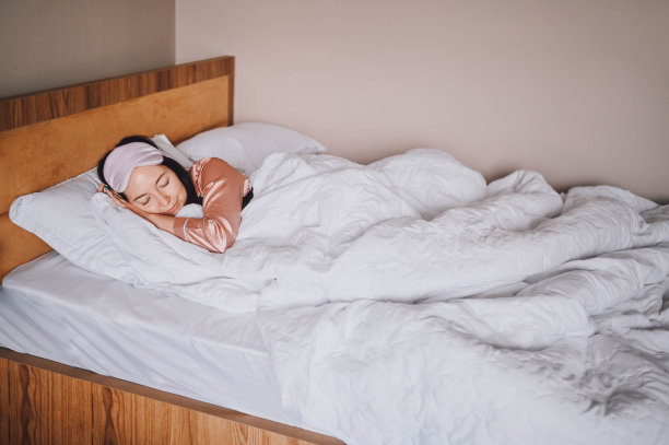 迷人微笑的年轻女子穿着粉红色的丝绸睡衣，带着眼罩在床上伸伸懒腰，独自醒来快乐的概念，醒来后健康的睡眠在舒适的床上和床垫享受早安。图片下载