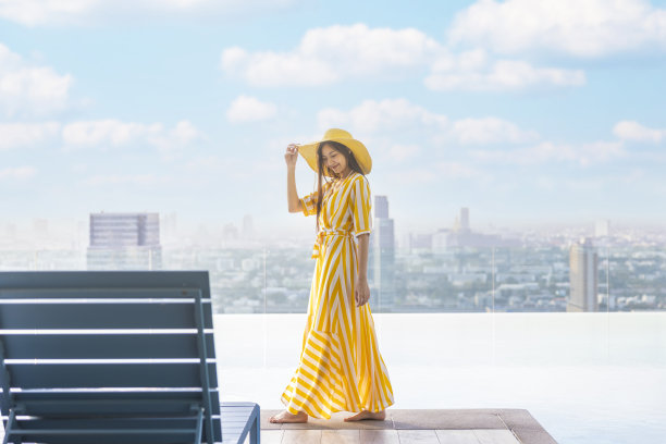 美丽的女人，穿着黄色的衣服，戴着黄色的帽子，走在无限的游泳池边，模糊的背景，现代的城市风景和美丽的天空与云彩。集中注意力，女士，复印空间图片下载