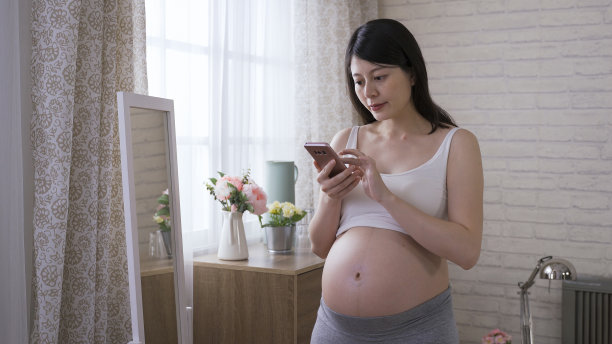 一名快乐的女子在家拍怀孕照，一边轻敲电话，一边对着镜子里的自己微笑。图片下载