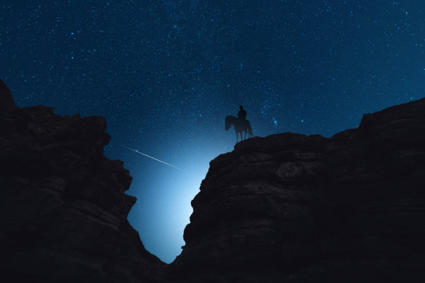 星夜里，独自骑马的人站在岩石上的剪影。他身后是美丽的夜空。图片下载