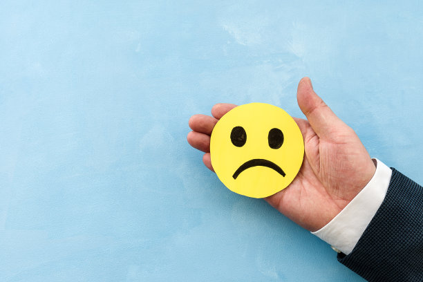 一个穿着西装的男人的手拿着一张黄色的笑脸，蓝色的背景上有一个不满的脸图标。顾客不满或负反馈的概念。图片下载