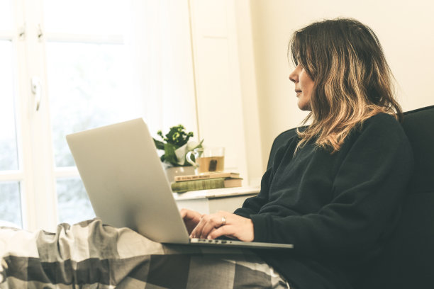 放松的女孩穿着暖和的睡衣坐在沙发上用笔记本电脑。早上，一个女人坐在客厅的沙发上用电脑工作。家庭工作、通信和技术概念。图片下载