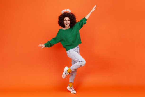 一名留着非洲式发型的快乐兴奋的女性，穿着绿色休闲风格的毛衣站在那里，抬起双臂，露出兴奋的表情。图片下载