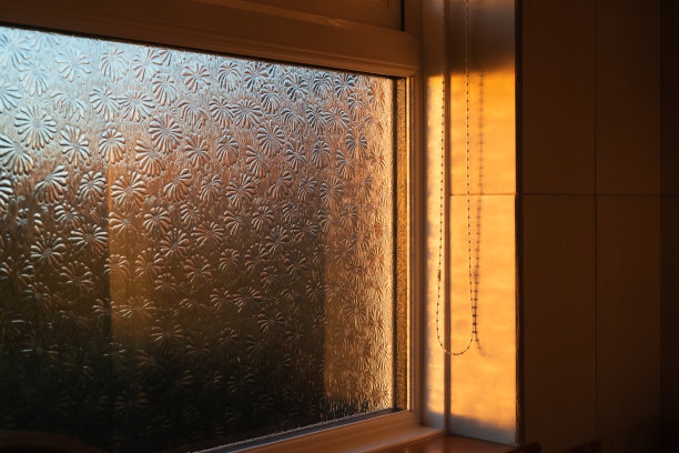浴室窗户上有图案的隐私玻璃，捕捉着夕阳的金色光线。可以看到用于卷帘的帘线垂下来。图片下载