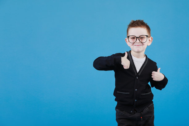 喜欢的。戴着眼镜的快乐小男生的肖像，对着镜头微笑，竖起大拇指，表示赞同。室内工作室拍摄孤立的蓝色背景图片下载