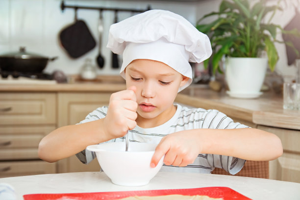 一个戴着白色厨师帽的白人男孩在厨房的餐桌上用勺子在碗里搅拌。小孩帮他妈妈做自制蛋糕。图片下载