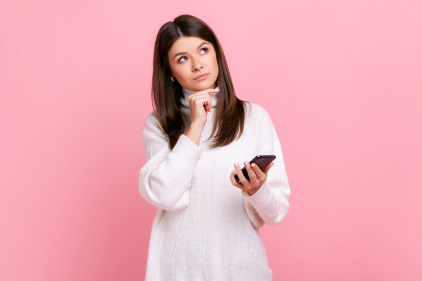 深思熟虑的漂亮年轻的黑发女孩手里拿着智能手机，看向别处，思考。图片下载