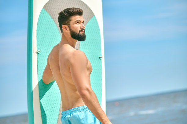 一个身材魁梧的年轻人在沙滩上拿着冲浪板图片下载