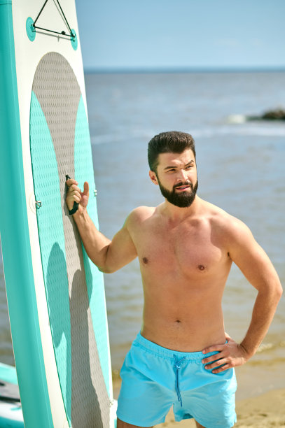 一个身材魁梧的年轻人在沙滩上拿着冲浪板图片下载