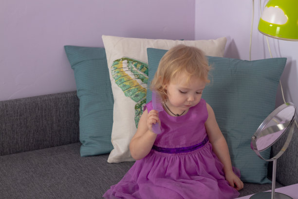 一个穿着漂亮紫色连衣裙的小女孩在她房间的沙发上用梳子梳头发图片下载