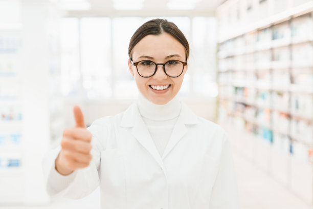 特写裁切聚焦镜头微笑的女性白人药剂师药剂师在药店的白色医疗大衣在相机里竖起大拇指图片下载