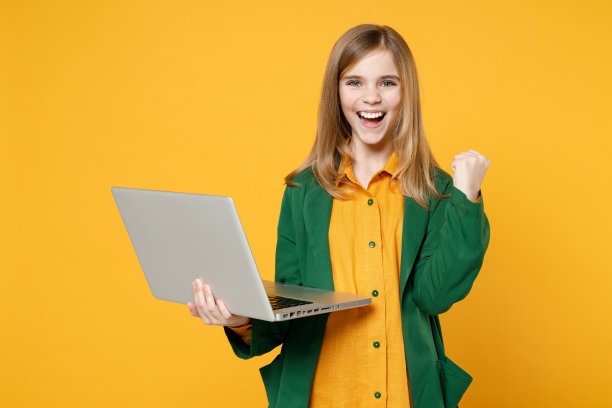 金发碧眼可爱可爱的小女孩，12-13岁，穿着休闲的衣服，拿着笔记本电脑，电脑，做胜利者的姿势，握紧拳头，孤立在黄色背景的儿童工作室。童年的生活方式的概念。图片下载