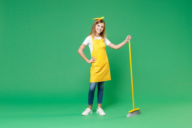 全身微笑的小女孩，家庭主妇12-13，围裙站着，手臂叉腰，拿着扫帚做家务，孤立在绿色背景的儿童工作室肖像。管家的概念。图片下载