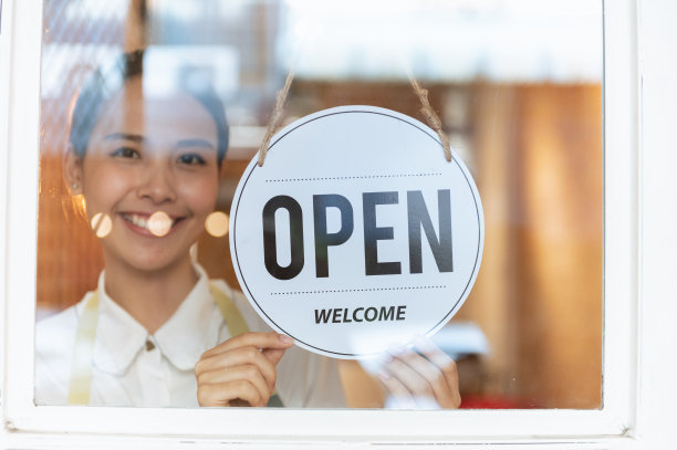 在因冠状病毒大流行而被隔离后，亚洲小企业主妇女把商店入口的招牌重新打开。一个女人在玻璃窗上挂着开着的牌子。关注的迹象。图片下载