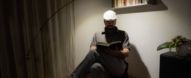 年轻人坐在家里，在他客厅角落的灯下的椅子上看书，放松和享受他的活动和自由时间。图片下载