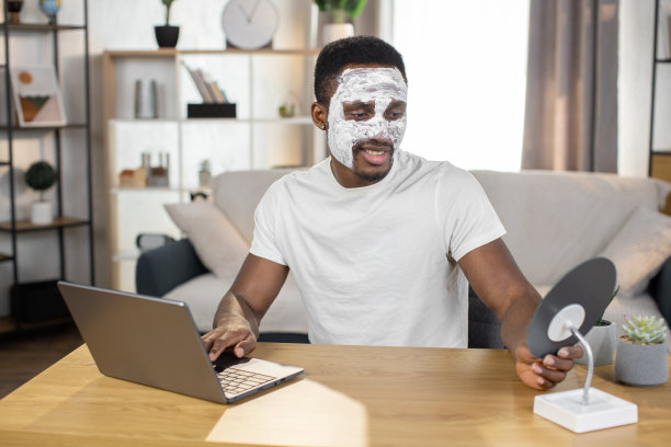 戴着白色面膜的非裔美国人，坐在家里的桌子上，在笔记本电脑上打字图片下载