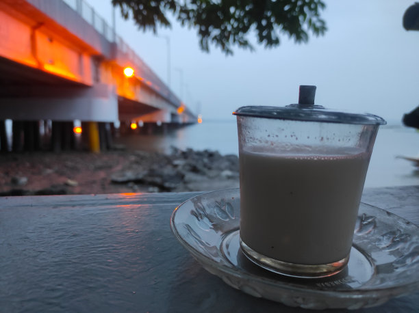 近景照片:桌上端着一杯热巧克力牛奶，落日余辉映衬着苏拉玛杜大桥下的海岸线。图片下载