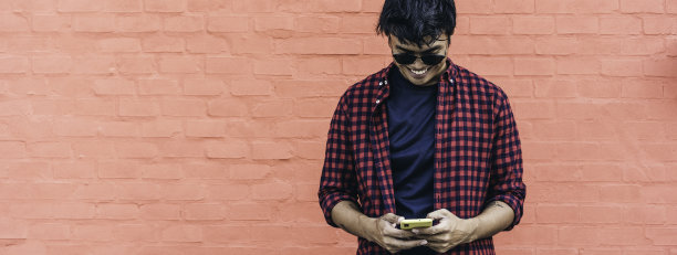 挂着面带微笑的亚洲年轻人在户外对着红墙使用手机的横幅——亚洲社会影响者在智能手机应用程序的新趋势中享受乐趣——千禧一代年轻人的生活方式图片下载