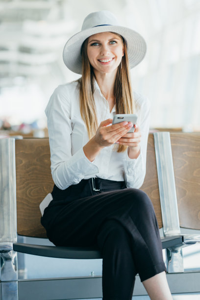 女孩在候机室用智能手机。美丽的年轻女孩使用智能手机通讯应用程序机场抵达新的国家。女人环游世界。对着镜头微笑图片下载