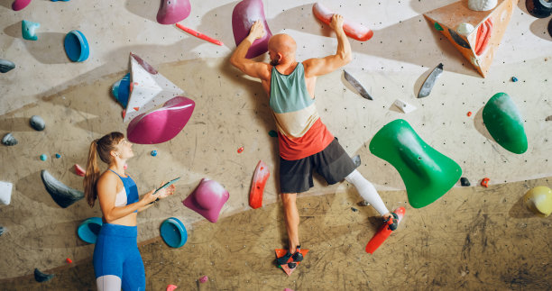 攀岩教练在健身房的抱石墙上给初学者上课。女运动员拿着平板电脑，向强壮的男运动员展示正确的攀岩技巧。图片下载