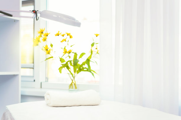 近距离的照片，一个美容师空的办公室与护理化妆品和按摩台与放大镜灯和黄色的天然花。图片下载
