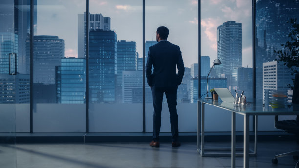 自信的年轻商人，穿着考究的西装，站在他的现代办公室里，看着窗外高楼林立的大城市。成功的财务经理计划项目策略。图片下载