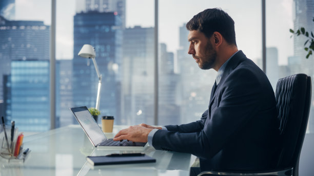 穿着西装的自信商人坐在现代办公室的办公桌前，使用笔记本电脑，紧靠窗外的大城市和摩天大楼。成功的财务经理计划工作项目。图片下载