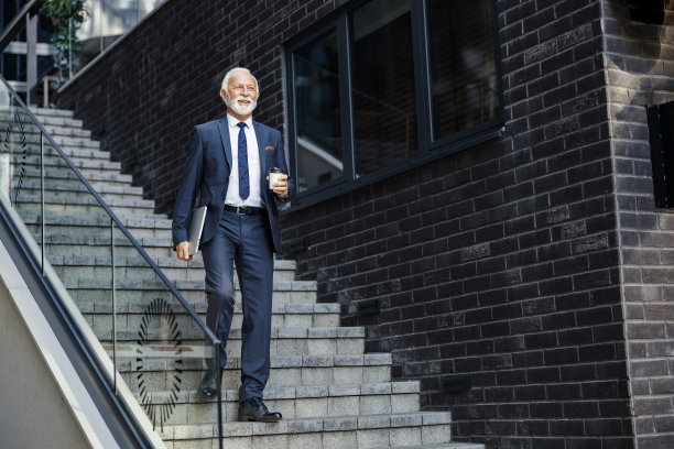 一位老经理下楼去他的办公室。一位快乐的高级商人在商务中心走下楼梯，腋下夹着一台笔记本电脑，喝着咖啡走进他的办公室图片下载