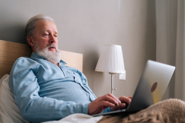 低角度的观点聚焦灰白头发的老年男性与胡子使用笔记本电脑躺在床上坐在家里。图片下载