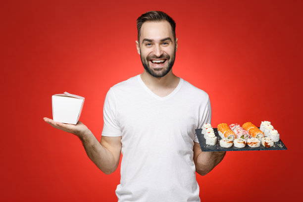 微笑的20多岁的年轻人，穿着基本的白色t恤，把中国菜装在外卖纸盒里，装在饭盒里，寿司卷装在黑色的盘子里，传统的日本食物被隔离在红色的背景上。图片下载