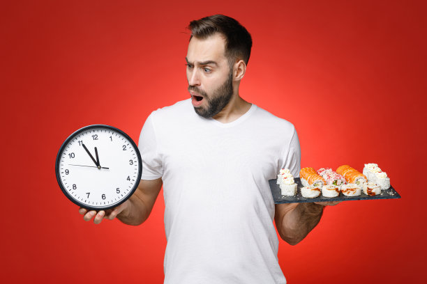 一名20多岁的大胡子青年穿着休闲的白色t恤，抱着钟，寿司卷装在黑色盘子上，传统的日本食物被隔离在红色的背景上，工作室肖像。图片下载