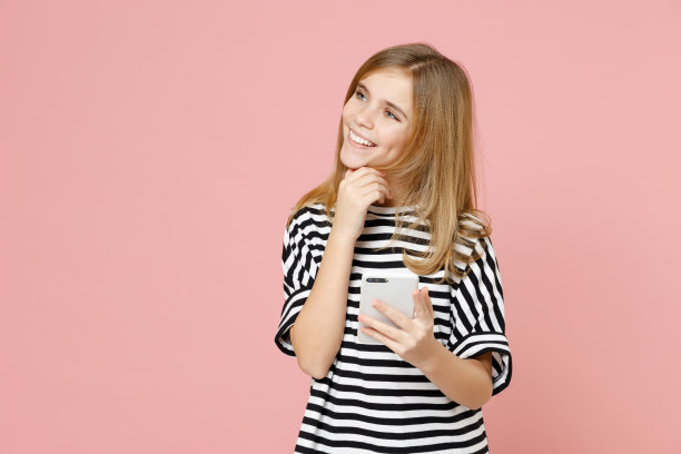 小金发微笑的小女孩12-13岁，穿着条纹的超大t恤，拿着手机，撑着下巴，看着一边孤立在淡粉色背景下的孩子。童年的生活概念图片下载