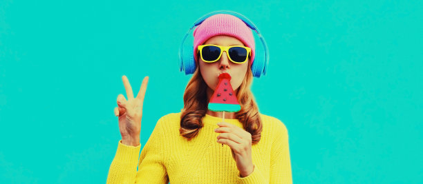夏天新鲜多彩的快乐年轻女子的肖像，戴着耳机听着音乐，水果多汁的棒棒糖或冰淇淋形状的西瓜片在蓝色的背景上图片下载