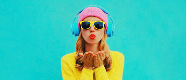 时尚年轻女子的肖像，戴着耳机听着音乐，吹着她的嘴唇送甜蜜的空吻，穿着彩色的黄色毛衣在蓝色的背景图片下载