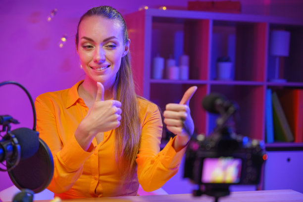 在一间紫色灯光下的摄影棚里，一位年轻可爱的女性博主，金发碧眼，身穿黄色衬衫，正对着摄像机，竖起大拇指图片下载