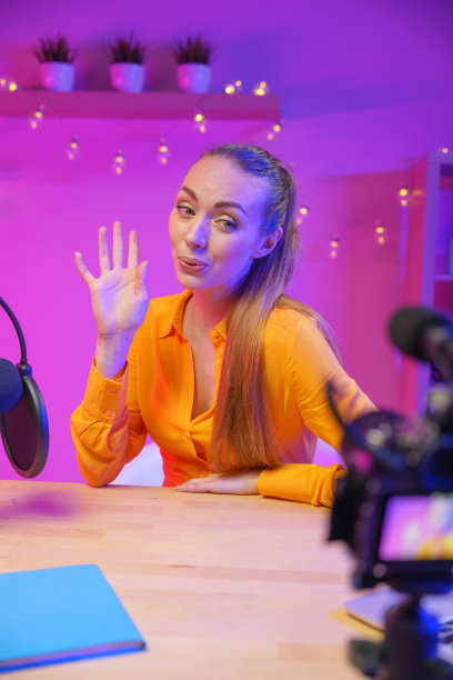 一位有魅力的年轻博主，金发碧眼，身穿黄色衬衫，在镜头前挥手告别，坐在一张桌子前，前台有一台摄像机图片下载