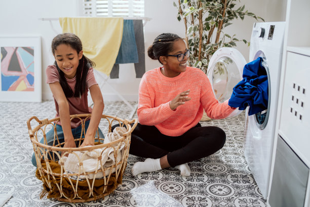 女孩和妈妈坐在洗衣房的地板上，星期六打扫房子，做家务，女儿从脏衣篮里拿出衣服扔到洗衣机里图片下载