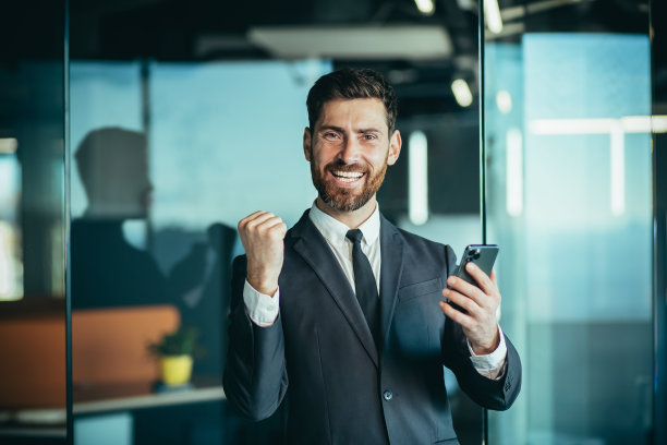 开心的商人老板看着相机微笑着，手里拿着手机，男人在网上赢了，庆祝胜利图片下载