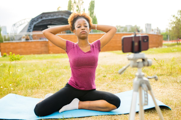 在夏日公园的户外，一名西班牙裔女性博主穿着运动服，在垫子上做瑜伽，用手机拍摄视频图片下载