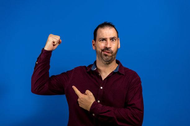 有趣的留着胡子的年轻人穿着蓝色背景的紫色衬衫。人们生活方式的概念。模拟复制空间。展示肱二头肌，做胜利者的姿势。图片下载