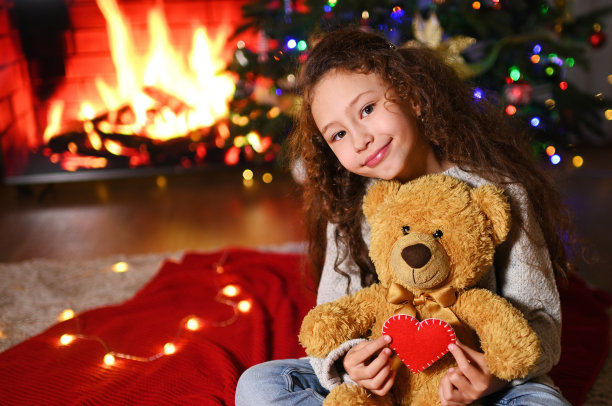 这个孩子是一个可爱的女孩，卷着头发，微笑着坐在一个红色的格子花呢上，拿着一个泰迪熊和一颗红心在壁炉的背景下图片下载