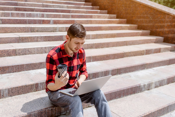 一个年轻的自由职业者正在户外用他的笔记本电脑工作，喝着咖啡图片下载