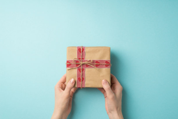 第一人称俯视图照片的情人节装饰女孩的手给予工艺纸礼品盒与心形图案丝带和缠绕蝴蝶结在孤立的淡蓝色背景与空白空间图片下载