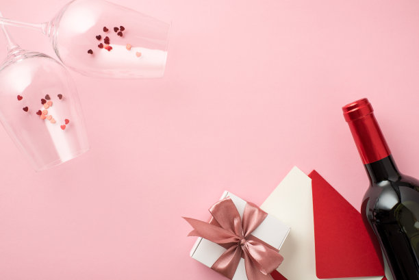 顶视图照片的saint valentine's day装饰葡萄酒杯与心形纸花白色礼盒粉红色蝴蝶结红色信封卡和酒瓶在孤立的淡粉色背景与copyspace图片下载