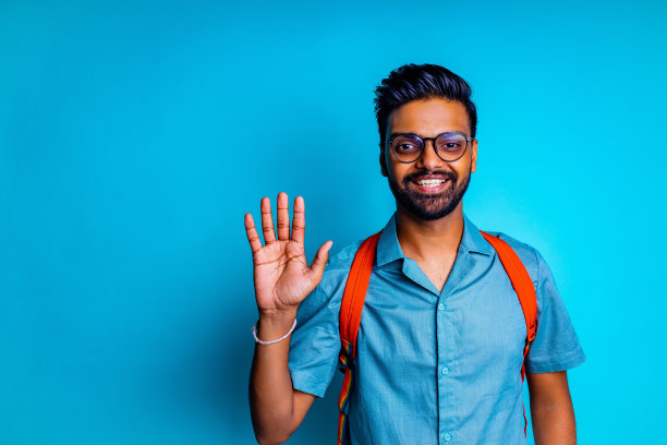一名印度学生戴着眼镜，背着橙色背包，在蓝色的画室里用手打招呼，看着相机感到很开心图片下载