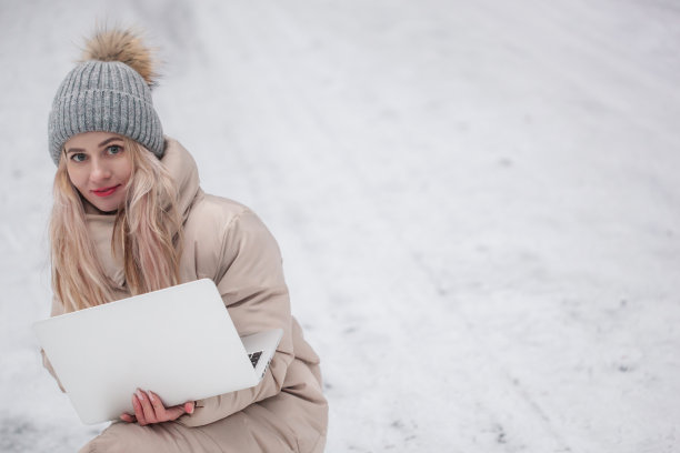 美丽的微笑的女孩和笔记本电脑坐在冬季公园工作。图片下载