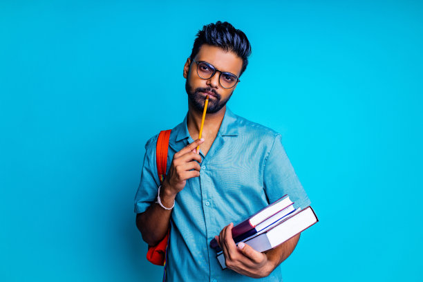 英俊的年轻印度男子，留着胡子，戴着眼镜，蓝色的棉t恤，橙色的彩虹背包，工作室背景里有书图片下载