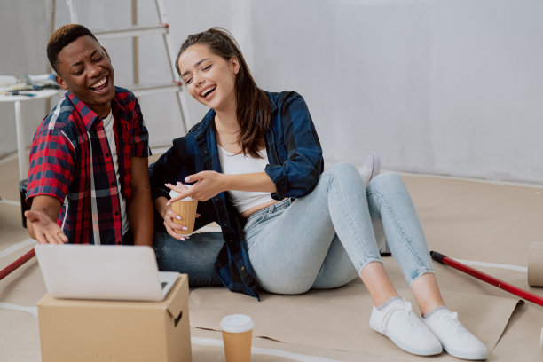 在装修过的公寓里，一对对相爱的情侣坐在铺满纸箱的地板上浏览着笔记本电脑，家具装饰，粉刷墙壁，休息一下，喝着咖啡准备带走图片下载