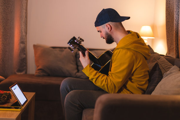 一个留着大胡子的年轻人一边在家里学习弹吉他，一边用他的iPad平板电脑在网上看教程和乐谱图片下载