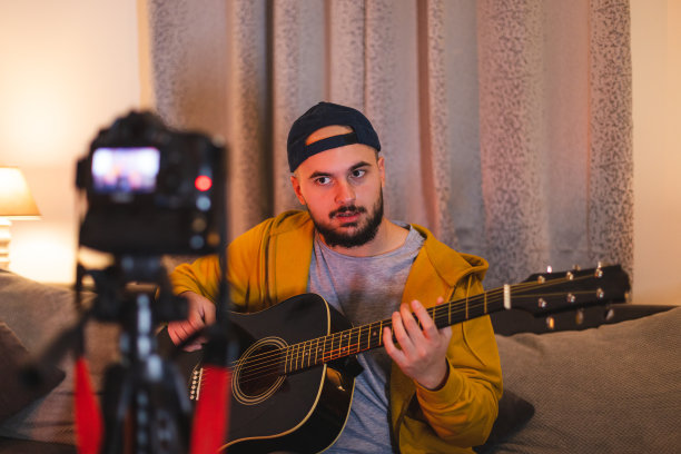 一个留着大胡子的年轻人一边在家里弹吉他，一边用单反相机为YouTube录制教程图片下载